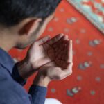 A young man holding his hands in prayer on a patterned mosque carpet, symbolizing devotion.