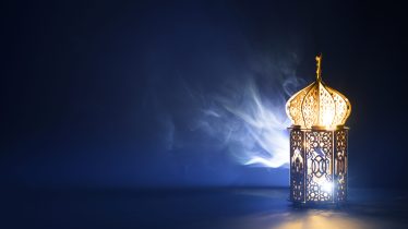 Ornamental Arabic lantern with burning candle glowing at night and glittering golden bokeh lights. Festive greeting card for Muslim holy month Ramadan Kareem.