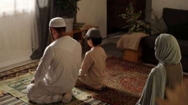 High angle view of unrecognizable Muslim family with kid sitting on floor in living room praying salah