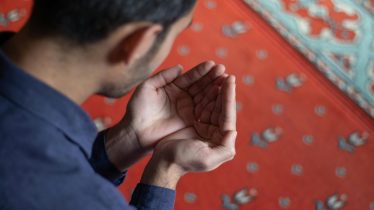 A young man holding his hands in prayer on a patterned mosque carpet, symbolizing devotion.