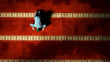 A single person in prayer on a beautifully patterned carpet in a mosque, viewed from above.